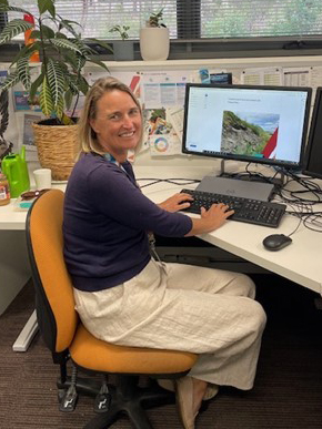 A woman sitting at a desk in an office. She has her hands on the keyboard and has turned to smile at the camera.