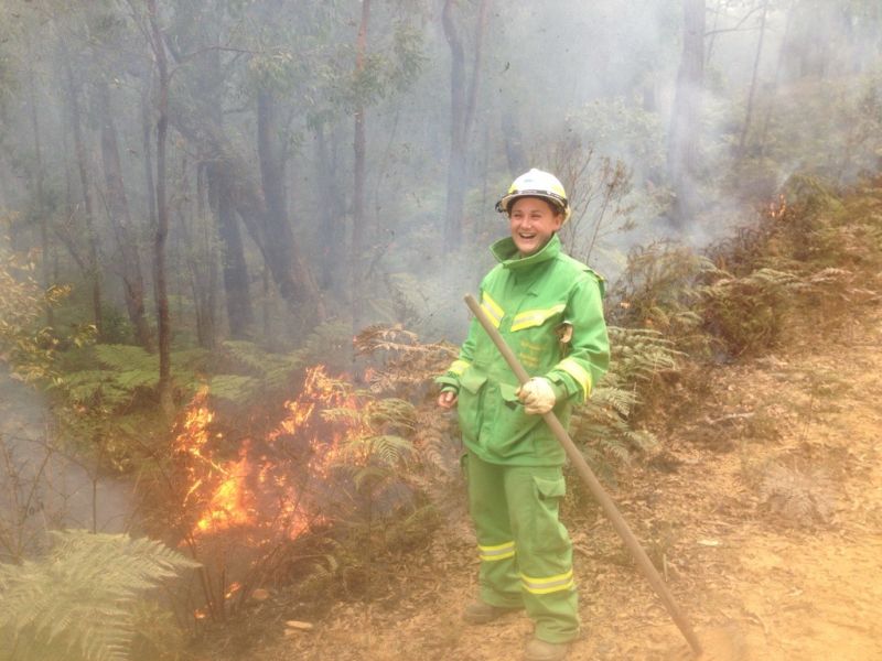 A female firefighter wearing a helmet and green overalls. She is holding a rake hoe and conducting a planned burn in bush scrub