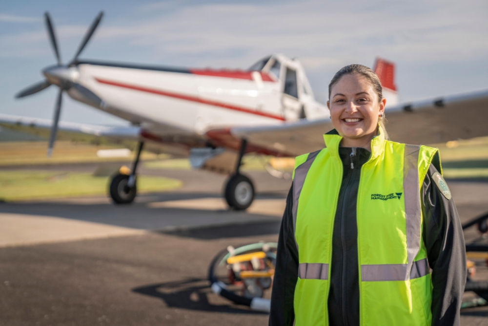 Woman smiles at camera wearing fluoro yellow vest, with small white plane in the background