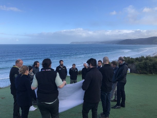 A group of people are gathered beside the ocean. They are standing in a circle, looking at a large poster.