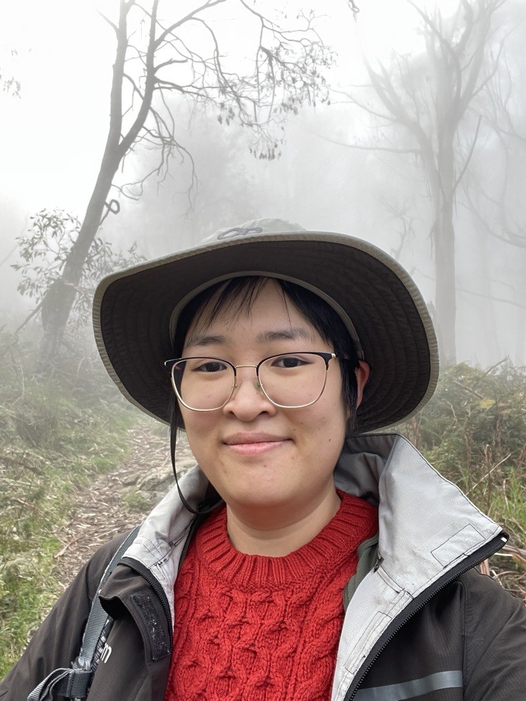 Photo of young woman, wearing glasses, a red knit top, an open coat. There are trees behind her and it's a foggy day