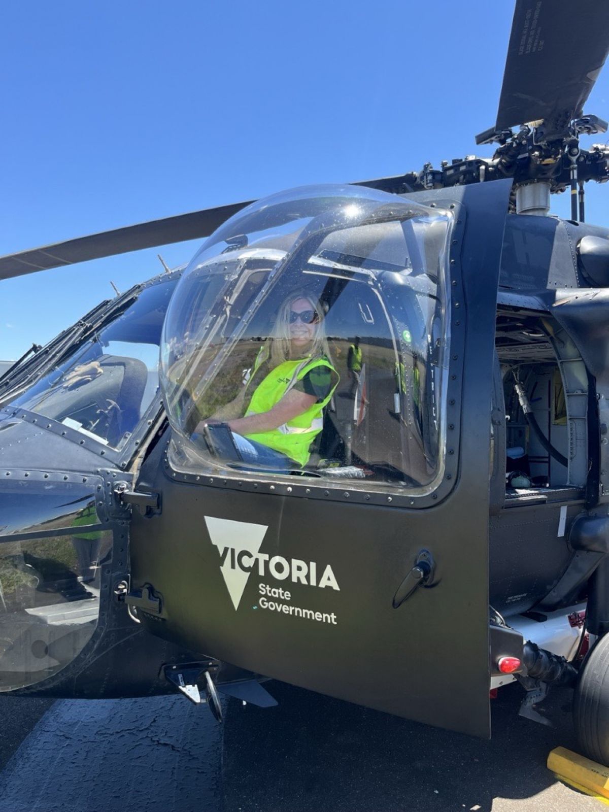 Sharon O'Sullivan sitting in landed Blackhawk Helicopter at Essendon Airport. The door is open and she is facing the camera, wearing sunglasses